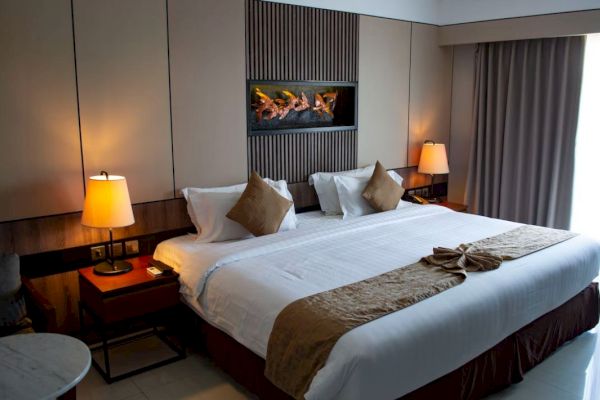 A modern hotel room with a large bed, throw pillows, two bedside lamps, a wall-mounted fireplace, and a curtain-covered window.