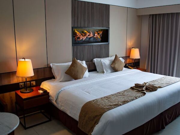 A modern hotel room with a large bed, throw pillows, two bedside lamps, a wall-mounted fireplace, and a curtain-covered window.