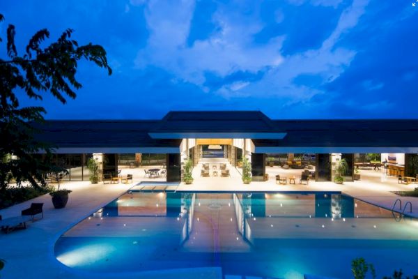 The image shows a luxurious, lit-up resort with a large swimming pool, outdoor seating, and a modern building against a blue evening sky.