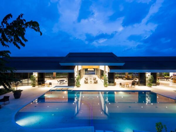 The image shows a luxurious, lit-up resort with a large swimming pool, outdoor seating, and a modern building against a blue evening sky.