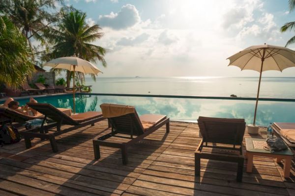 Poolside deck with lounge chairs and umbrellas overlooking a calm sea under a partly cloudy sky, surrounded by palm trees and relaxing vibes.