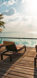 A serene poolside scene with sun loungers, umbrellas, palm trees, and a view of a calm ocean under a partly cloudy sky, during sunset or sunrise.