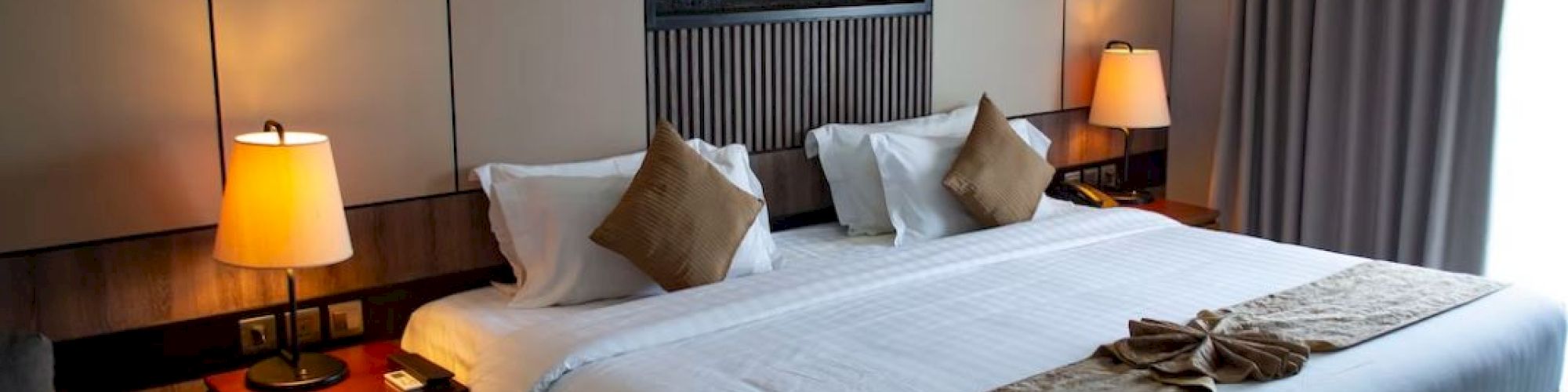 This image shows a modern hotel room with a neatly made king-sized bed, two bedside tables with lamps, and a window covered by curtains.