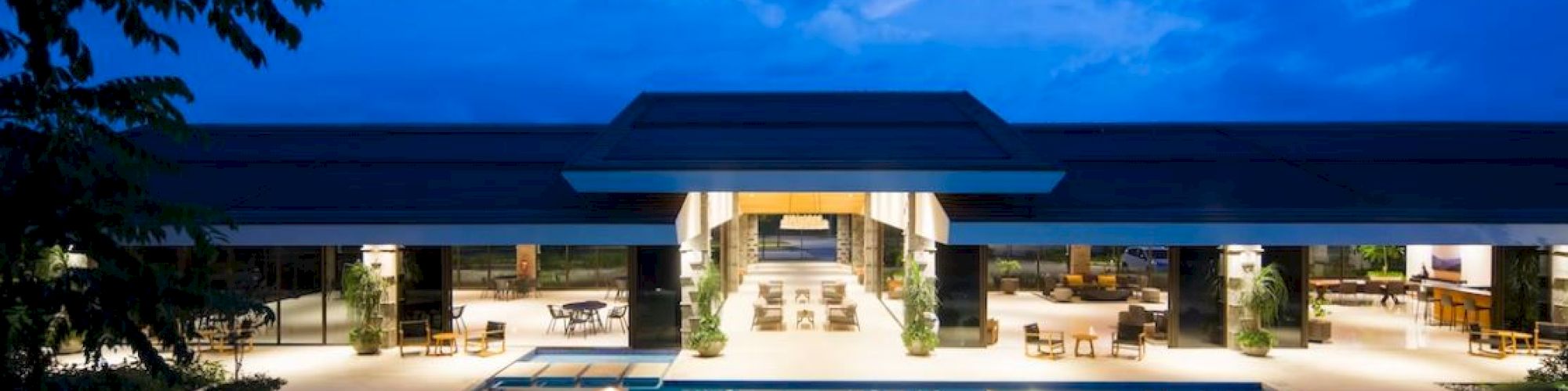 A luxurious, modern building with a large, illuminated swimming pool in the forefront, set against a blue sky backdrop during twilight.