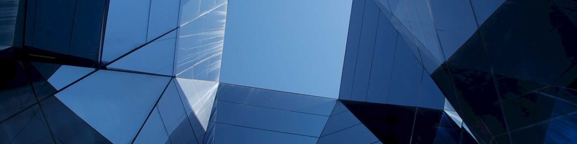 The image shows a series of modern glass buildings or structures reflecting the sky, creating a geometric pattern with a central opening.