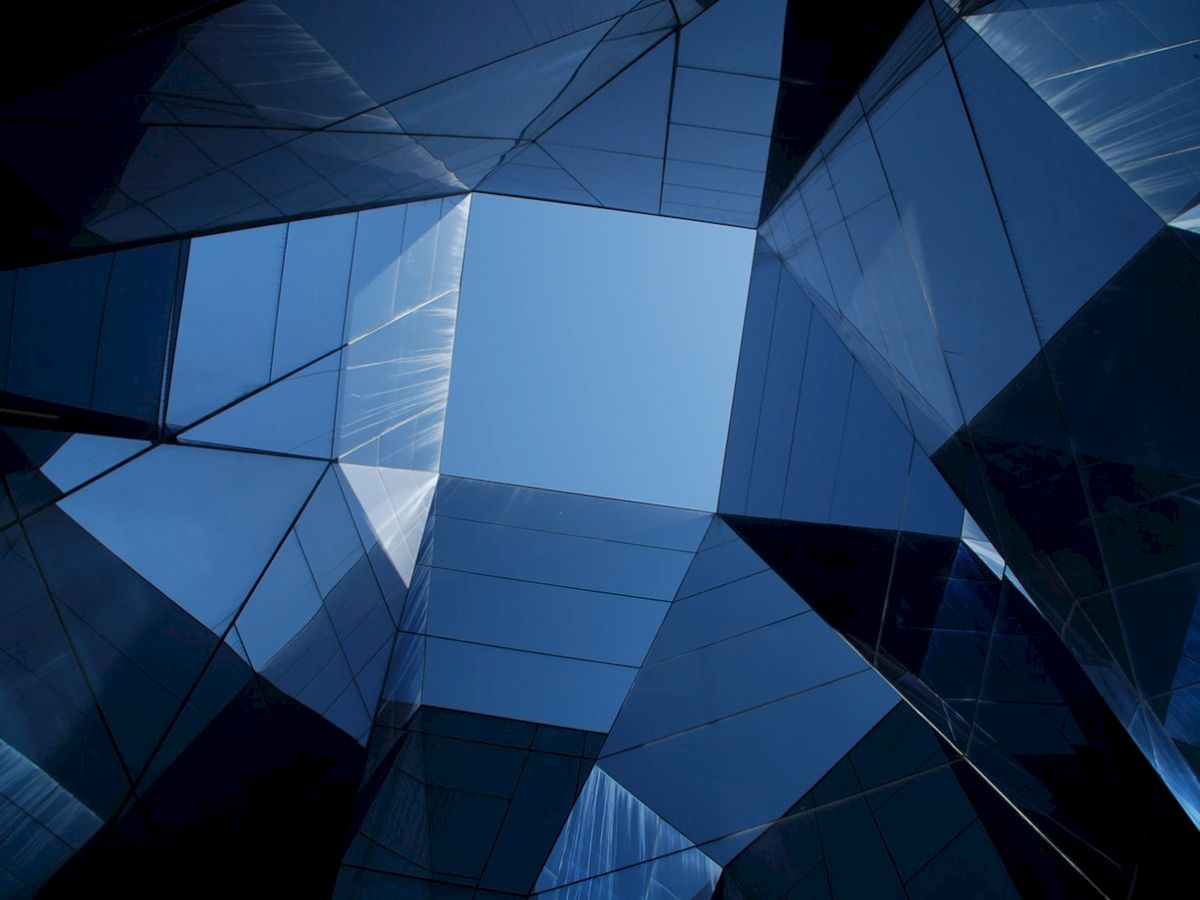 The image shows an upward view of modern, reflective glass buildings, forming a geometric pattern with a clear blue sky in the center.