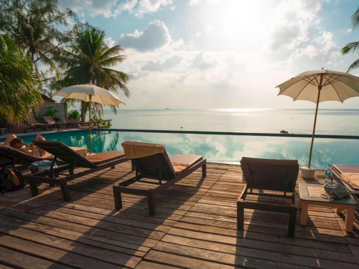 This image shows a serene poolside scene with lounge chairs, umbrellas, palm trees, and a view of the ocean under a partly cloudy sky.