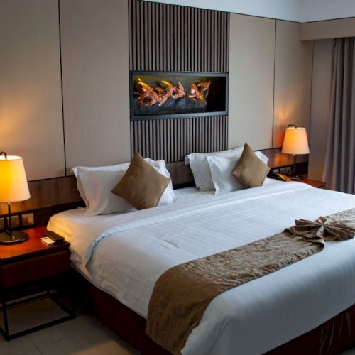 A neatly decorated hotel room features a king-size bed with a headboard, two bedside tables, lamps, and a wood wall accent.