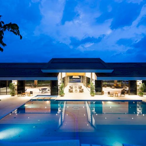An illuminated modern building with a large outdoor pool at night, featuring lounge areas and surrounded by trees under a cloudy blue sky.