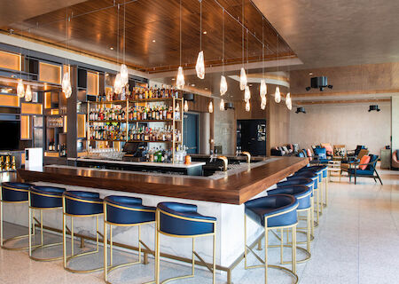 A modern, stylish bar with blue stools, hanging lights, and a stocked bar area; a cozy seating area is visible in the background.