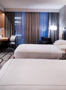 A modern hotel room with two beds, a wall-mounted TV, a desk with a chair by the window, and stylish decor.