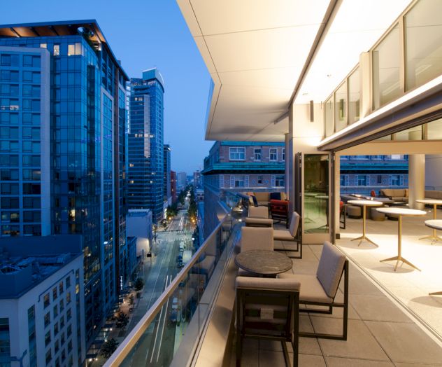 A modern cityscape at dusk with high-rise buildings and an elegant open-air balcony featuring seating and tables on the right side.