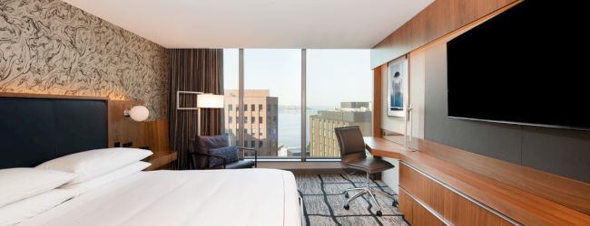A modern hotel room featuring a large bed, desk, chair, TV, and a floor-to-ceiling window with a city and water view in the background.