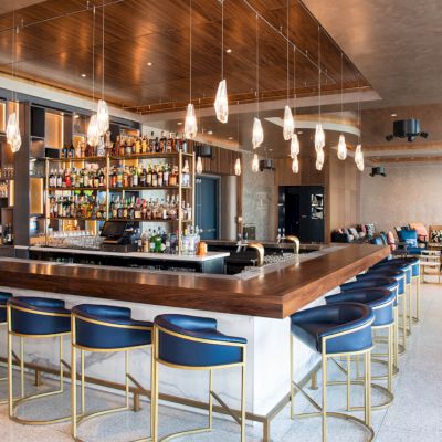 This image shows a modern, upscale bar with a wooden countertop surrounded by blue chairs, hanging lights, and a fully stocked back bar.