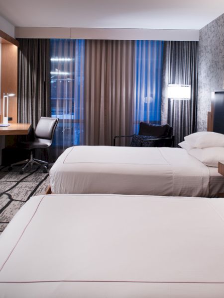 The image shows a modern hotel room with two neatly made beds, a wall-mounted TV, a desk, and a chair near the window with curtains.
