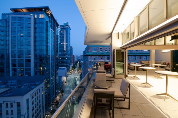 A modern cityscape at dusk featuring tall buildings and a lit outdoor balcony with tables and seating on an upper floor.
