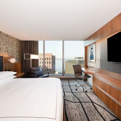 A modern hotel room with a large bed, desk, chair, and floor-to-ceiling window offering a cityscape view, featuring minimalistic decor.