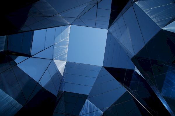 This image shows a view looking up at the sky through an arrangement of modern glass buildings, creating geometric reflections and patterns.