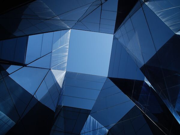 This image shows a view looking up at the sky through an arrangement of modern glass buildings, creating geometric reflections and patterns.