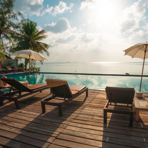 Several lounge chairs and umbrellas by a pool overlooking a calm ocean under a sunny sky, with trees in the background, creating a relaxing scene.