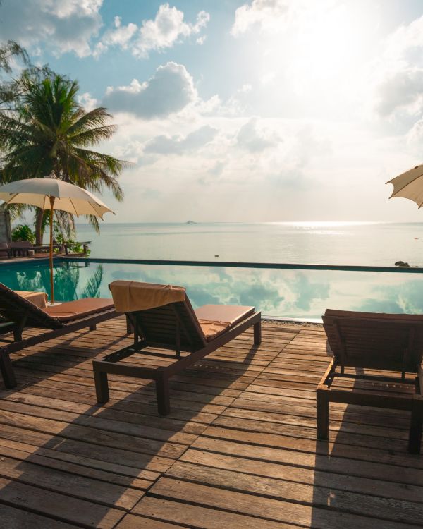 Several lounge chairs and umbrellas by a pool overlooking a calm ocean under a sunny sky, with trees in the background, creating a relaxing scene.