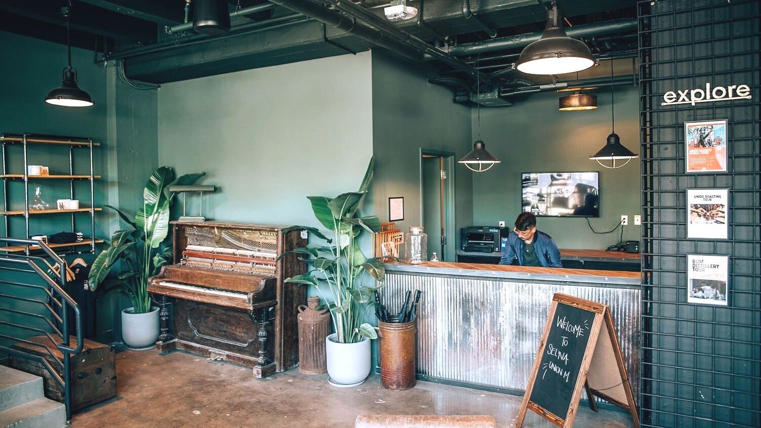 A modern reception area with a piano, plants, and decor. A person is behind the front desk, with a chalkboard sign and industrial lighting.