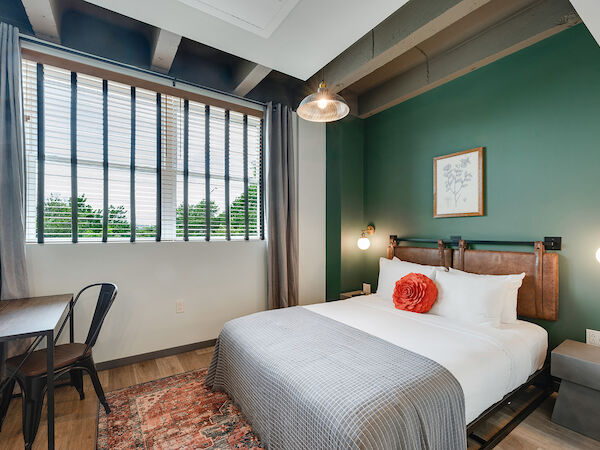 A cozy bedroom with a modern aesthetic features a bed with white sheets, a gray blanket, a red pillow, a wooden desk, and a large window.