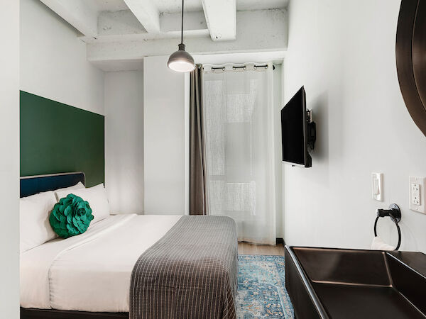 A modern bedroom with a neatly made bed, wall-mounted TV, round mirror, pendant light, and a sink. The room is decorated in a minimalist style.