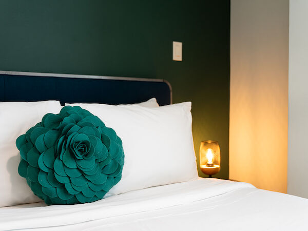 A neatly made bed with white linens features a green decorative pillow shaped like a flower and a lit bedside lamp on a dark background.