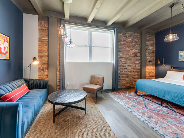 A modern bedroom with exposed brick walls, blue sofa, round coffee table, armchair, king-size bed, rug, art on walls, and hanging lights.