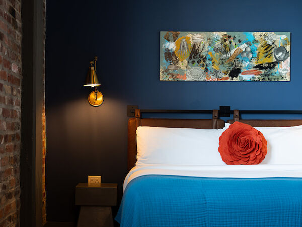 A neatly made bed with a blue blanket, a red flower cushion, brick wall, wall art, and a bedside table with a lamp in a cozy room.