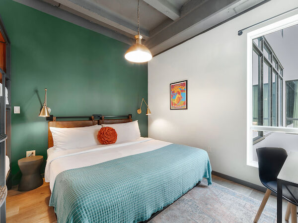 A modern bedroom with a green accent wall, a bed with a teal blanket, hanging lights, a framed picture, and a window with a view of an adjacent structure.