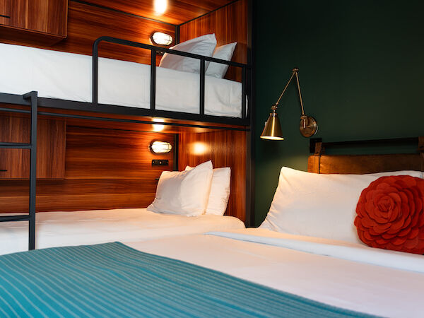 The image shows a cozy room with a bunk bed and a double bed, featuring white linens, a red decorative pillow, a blue throw blanket, and warm lighting.