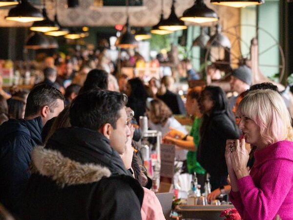 A crowd of people is gathered in a busy indoor market or food hall, engaging in conversations and transactions under hanging lights.
