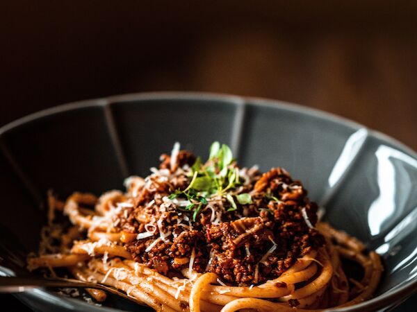 A hand holding a black bowl of spaghetti topped with a meat sauce and green garnish, set against a dark, out-of-focus background.