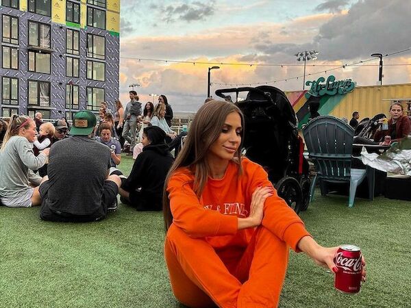 A woman in an orange tracksuit sits on the grass, holding a can of Coca-Cola, with a group of people and a tall building in the background.