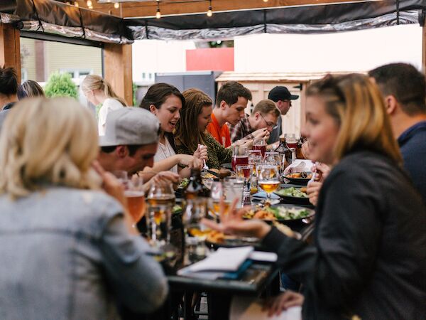 A group of people are gathered around a table, enjoying food and drinks at an outdoor setting, suggesting a casual gathering or social event.