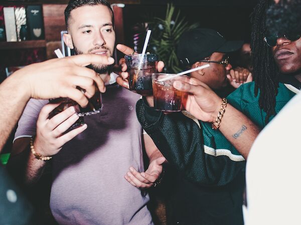 A group of people in a bar or club are raising their drinks for a toast, with a lively and social atmosphere in the background.