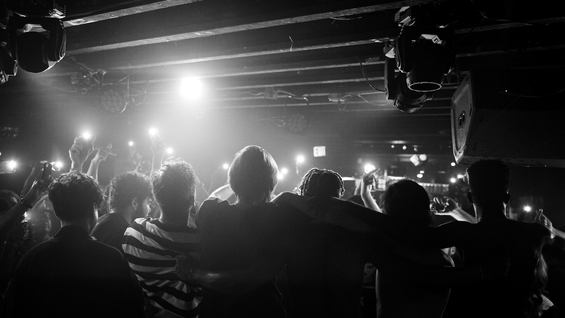 A group of people with their backs to the camera embrace under bright stage lights, holding up their phones to capture the moment.
