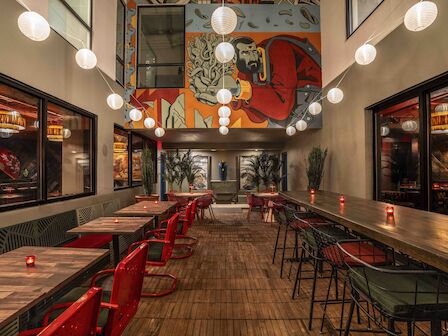 A stylish, industrial-chic restaurant interior with long wooden tables, red and green chairs, string lights, and a colorful mural at the back.