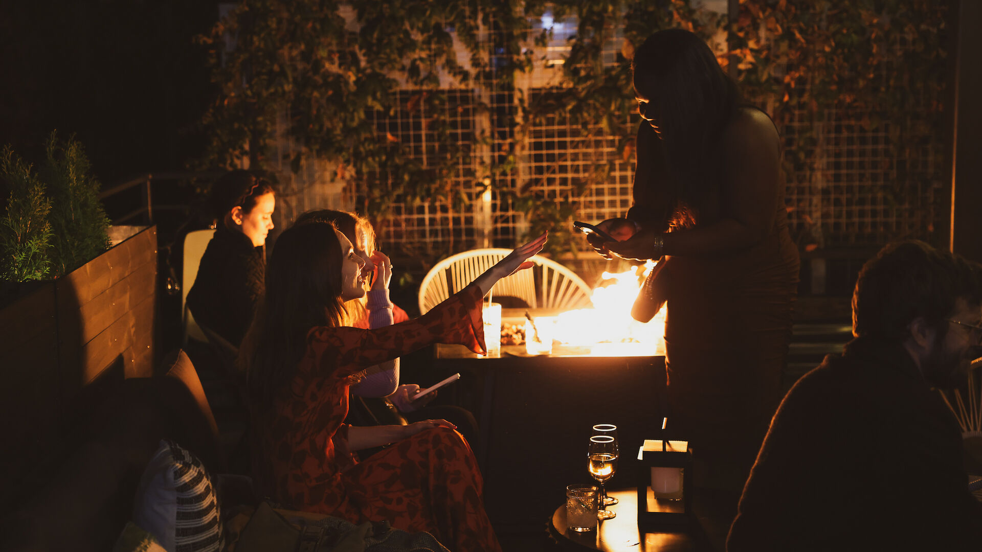 People sitting around a fire pit during what seems to be an evening gathering. Warm lighting illuminates the scene, with drinks on the table.