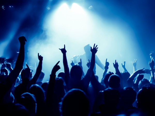 A crowd at a concert, cheering with hands raised, illuminated by vibrant blue stage lights and a haze of smoke.