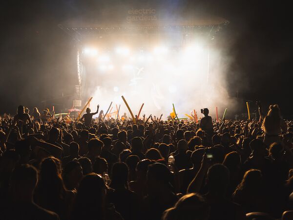 A large, energetic crowd is gathered at an outdoor concert at night, with people raising their hands and glow sticks under stage lights.
