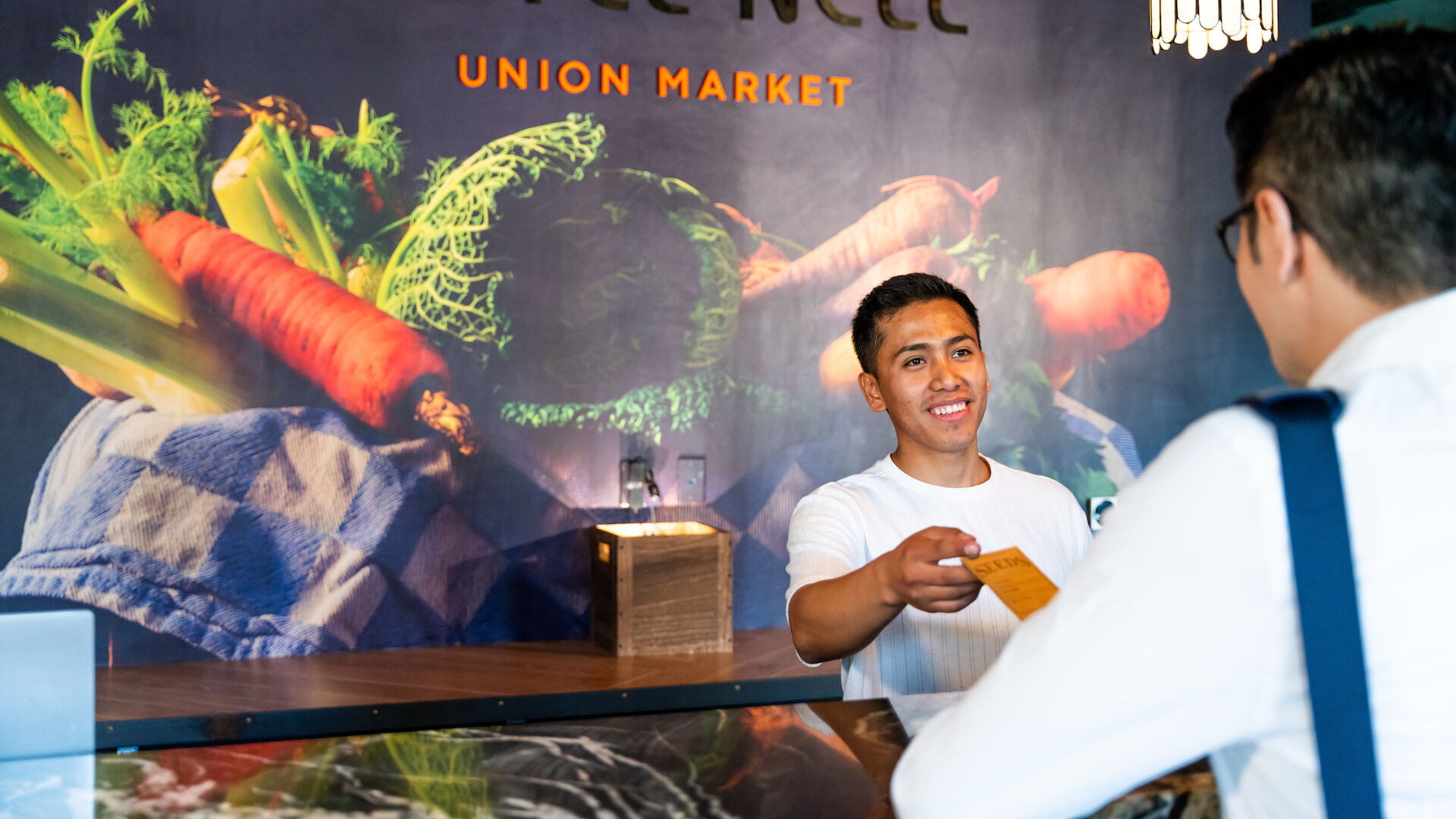 A front desk staff member at Hotel Nell's Union Market is handing a key card to a guest, with a colorful vegetable-themed background mural.