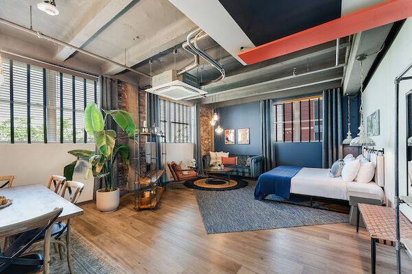 A modern, open-plan living space with a bed, seating area, dining table, and large plants, featuring exposed ceilings and bright decor.