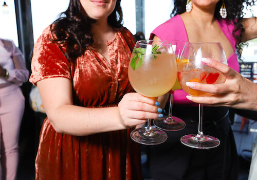 Three women holding and clinking glasses with various drinks, dressed in colorful outfits, at what appears to be a social gathering.