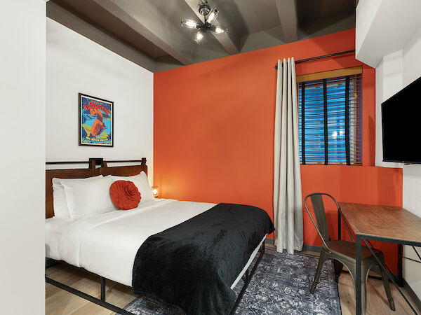 A cozy bedroom with an orange accent wall, a bed with white linens and a black throw, a desk with a chair, a wall-mounted TV, and a painting.