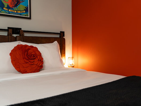 A well-made bed with white linens, a large orange flower pillow, an orange accent wall, a small lamp, and a framed poster on the wall.