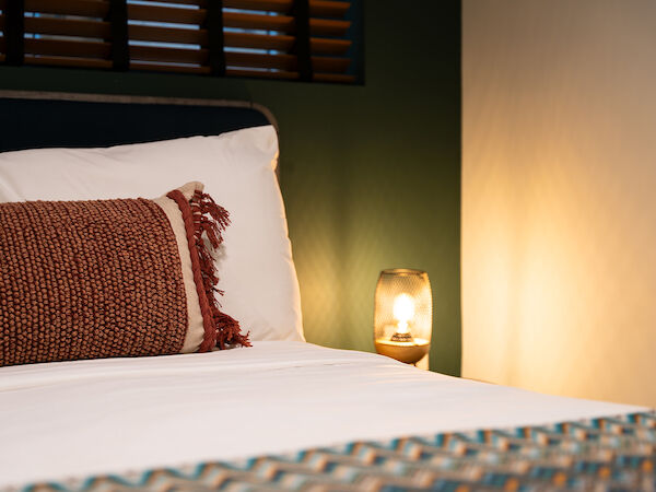 A cozy bedroom with a bed, a patterned throw pillow, a bedside lamp, white bedsheets, and a window with wooden shutters in the background.