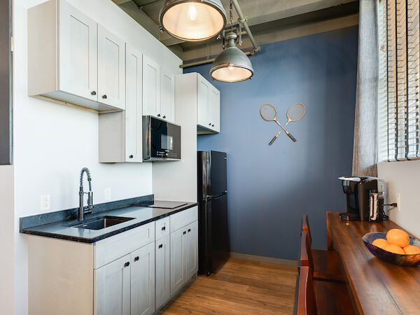 A modern kitchen with white cabinets, black countertops, a black fridge, and a microwave, features wall decor of two tennis rackets crossed.